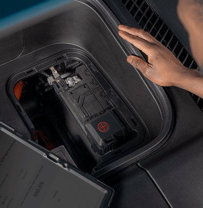 Nahaufnahme der Hand eines Technikers an einem geöffneten Fach, in dem die 12-Volt-Batterie und elektrische Komponenten zu sehen sind. Im Vordergrund zeigt ein Diagnosetablet den Ladezustand der Batterie in Prozent an.
